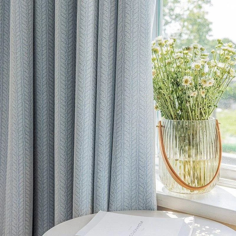 Rideaux élégants de couleur bleu clair avec un motif discret, suspendus près d'une fenêtre baignée de lumière naturelle. Un vase transparent contenant un bouquet de fleurs blanches est posé sur une table ronde, ajoutant une touche de fraîcheur et de délicatesse à la décoration d'intérieur.