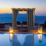 Pergola de terrasse élégante avec des rideaux foncés, située au bord d'une piscine sur une terrasse offrant une vue panoramique sur la mer au coucher du soleil. Ambiance relaxante et sophistiquée, idéale pour des soirées en plein air ou des moments de détente. Aménagement extérieur moderne parfait pour les maisons en bord de mer ou les espaces de loisirs en plein air.