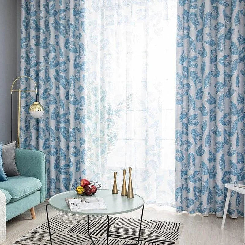 Rideaux à motifs de feuilles bleues dans un salon moderne, avec une lumière naturelle généreuse. Décoration contemporaine, idéale pour ajouter une touche de fraîcheur à votre intérieur. Ambiance relaxante avec un canapé bleu, une table basse design et des objets décoratifs élégants.
