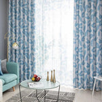 Rideaux à motifs de feuilles bleues dans un salon moderne, avec une lumière naturelle généreuse. Décoration contemporaine, idéale pour ajouter une touche de fraîcheur à votre intérieur. Ambiance relaxante avec un canapé bleu, une table basse design et des objets décoratifs élégants.