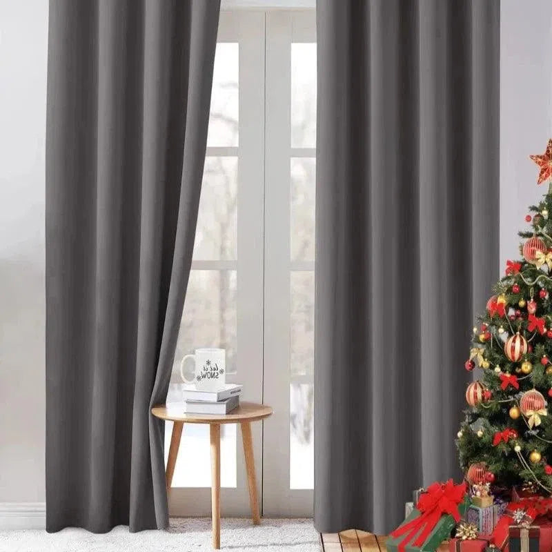 Rideaux opaques gris modernes pour salon, installés devant une grande fenêtre vitrée offrant une vue sur l'extérieur enneigé. Ambiance chaleureuse de Noël avec un sapin décoré et une petite table d'appoint accueillant une tasse et des livres, soulignant l'élégance de la pièce. Idéal pour créer une atmosphère cocooning et festive.