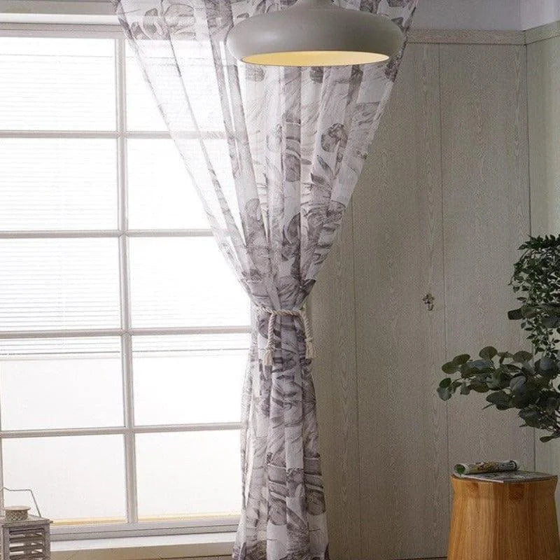 Rideau transparent à motifs floraux gris pour décoration intérieure, suspendu devant une grande fenêtre dans un salon moderne et lumineux. Design élégant avec des motifs subtils apportant une ambiance apaisante. Parfait pour ajouter une touche de style et de lumière naturelle à votre espace de vie tout en maintenant l'intimité. Adapté aux salons, chambres ou espaces de détente.