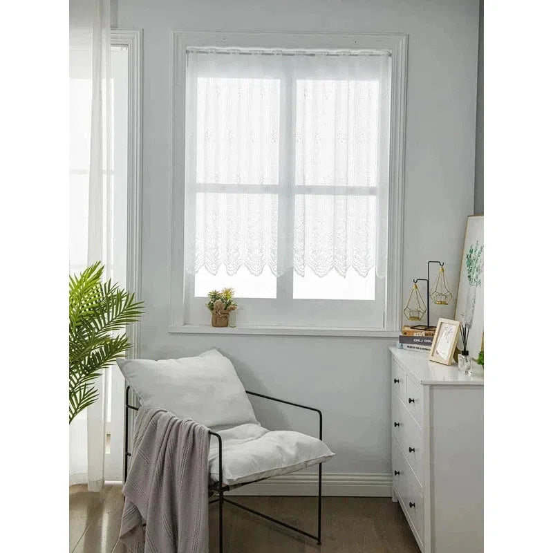 Décoration intérieure élégante avec rideau blanc dans un salon lumineux. La fenêtre est habillée d'un rideau léger, ajoutant une touche de sophistication à la pièce. Un fauteuil confortable avec un coussin est placé près de la fenêtre, devant une commode blanche décorée de cadres et de plantes. L'ambiance est épurée et moderne, parfaite pour créer un espace de détente dans la maison.
