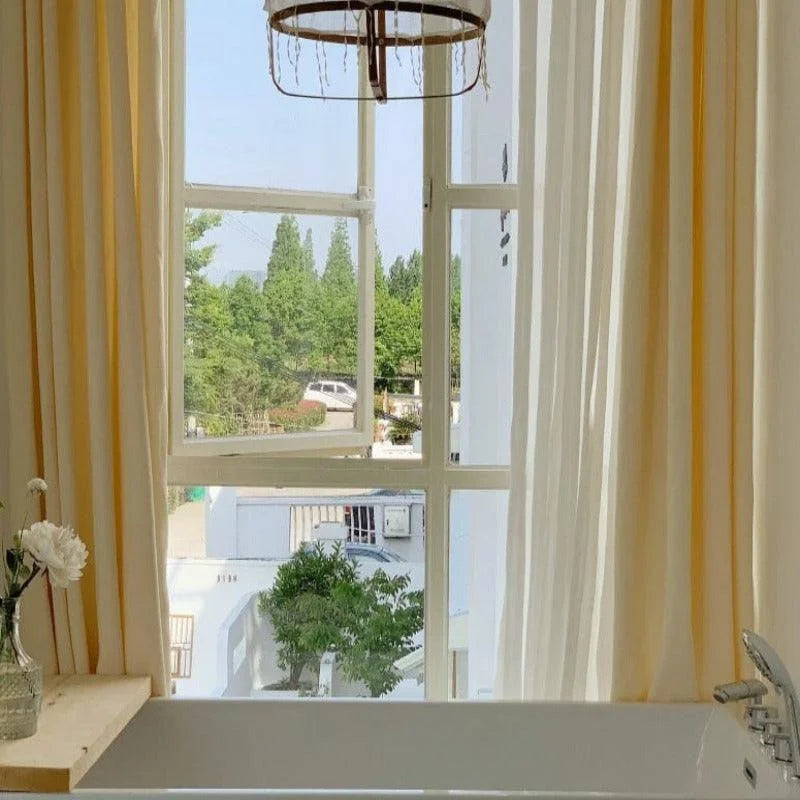 Baie vitrée lumineuse avec rideaux élégants laissant entrer la lumière naturelle, créant une atmosphère apaisante dans une salle de bain moderne. Un vase avec des fleurs rehausse la décoration, ajoutant une touche de fraîcheur et de nature. Vue extérieure verdoyante offrant un cadre relaxant et harmonieux. Robinet chromé visible sur le coin d'une baignoire, suggérant un espace de détente.