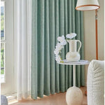 Rideau élégant de couleur bleu-vert, installé dans un salon moderne et lumineux. À côté, une table blanche décorative avec un vase rempli de fleurs blanches, ajoutant une touche florale et raffinée à la pièce. Une lampe sur pied avec un design contemporain complète le décor harmonieux.