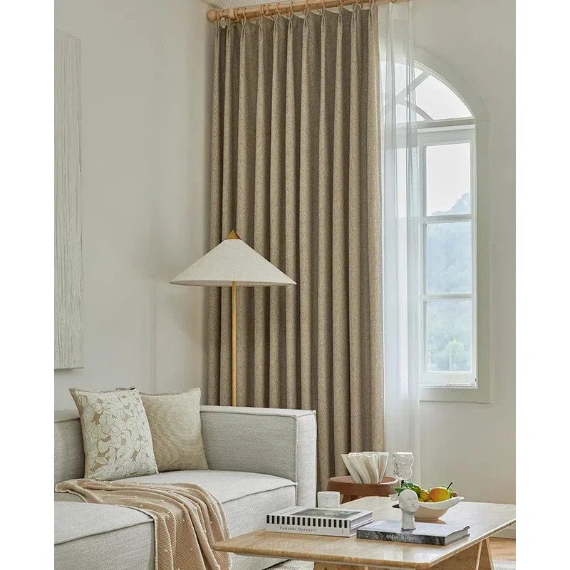 Canapé d'angle élégant et spacieux dans un salon moderne, accompagné de coussins décoratifs et d'une lampe sur pied. Rideaux longs à plis parfaits encadrant une fenêtre arquée laissant entrer la lumière naturelle. Table basse design en bois avec livres et décoration raffinée, créant une ambiance chaleureuse et accueillante. Idéal pour un intérieur contemporain et confortable.