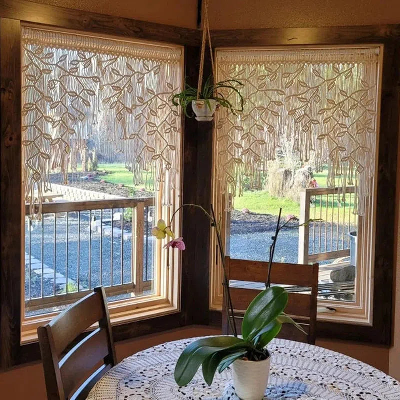 Rideaux déco style bohème avec motif en macramé ornant des fenêtres, créant une ambiance chaleureuse dans une salle à manger lumineuse. Une table ronde est agrémentée d'une nappe crochetée, avec une élégante orchidée en centre de table, offrant une touche de nature dans un espace intérieur accueillant. Vue sur un jardin en arrière-plan, idéal pour un aménagement intérieur harmonieux et tendance.