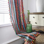 Décoration intérieure colorée avec rideau à motifs ethniques et pompons multicolores. Accent décoratif vibrant pour salon moderne. Vase en verre texturé vert avec plantes près d'une commode en bois clair. Idéal pour créer une ambiance bohème et chaleureuse.