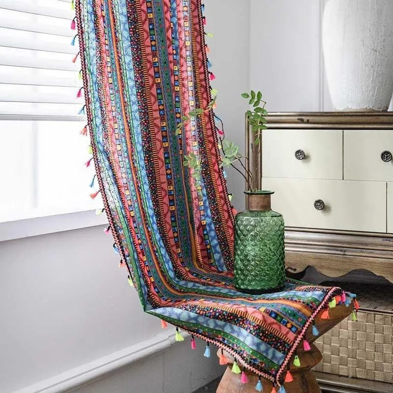 Décoration intérieure colorée avec rideau à motifs ethniques et pompons multicolores. Accent décoratif vibrant pour salon moderne. Vase en verre texturé vert avec plantes près d'une commode en bois clair. Idéal pour créer une ambiance bohème et chaleureuse.
