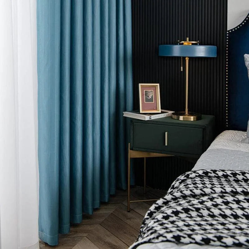 Chambre moderne avec rideaux bleus élégants, table de chevet sombre, lampe design et cadre photo, offrant une ambiance chic et contemporaine. Le motif du couvre-lit contraste avec les couleurs sombres, ajoutant une touche de sophistication à cet espace confortable et tendance.