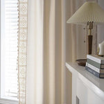 Rideau élégant beige pour salon, ajoutant une touche de sophistication à votre intérieur moderne, harmonisé avec une lampe de table et des livres disposés sur une cheminée blanche. Idéal pour créer une ambiance chaleureuse et accueillante dans votre maison.