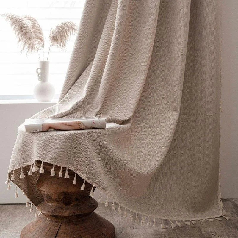 Rideau élégant beige avec franges, suspendu dans un intérieur moderne et minimaliste. Décoration de fenêtre apportant une touche cosy et chaleureuse à la pièce. Parfait pour créer une ambiance douce et naturelle dans votre maison. Adapté à tous types de décoration intérieure, soulignant la lumière naturelle filtrant subtilement à travers.
