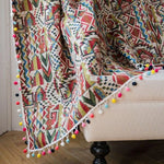 Jeté de canapé avec motifs géométriques colorés et pompons, relooking pour salon moderne et tendance, accessoire déco multicouleur pour une touche bohème.