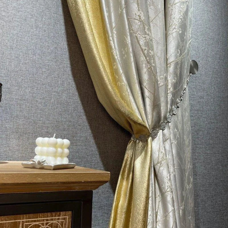 Rideau élégant beige et doré avec motifs floraux, accompagné d'un embrasse ornée de cristaux, accentuant le décor intérieur sophistiqué; une bougie décorative blanche posée sur une table en bois complète l'ensemble pour un intérieur chic et chaleureux.