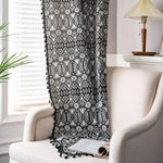 Plaid noir et blanc avec motifs géométriques, décoré de franges, posé sur un fauteuil beige dans un salon lumineux. Idéal pour ajouter une touche contemporaine à votre intérieur tout en apportant confort et chaleur. Parfait pour les soirées cocooning ou comme élément de décoration esthétique.