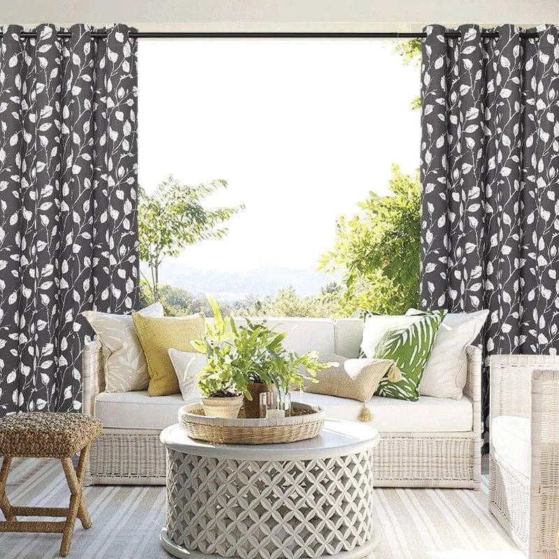 Rideaux à motifs feuillagés noirs et blancs encadrant une grande baie vitrée, laissant entrer la lumière naturelle dans un salon élégant. L'espace est aménagé avec un canapé confortable orné de coussins colorés et une table basse en rotin, créant une ambiance sereine et accueillante. Idéal pour un intérieur moderne et harmonieux.