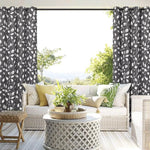 Rideaux à motifs feuillagés noirs et blancs encadrant une grande baie vitrée, laissant entrer la lumière naturelle dans un salon élégant. L'espace est aménagé avec un canapé confortable orné de coussins colorés et une table basse en rotin, créant une ambiance sereine et accueillante. Idéal pour un intérieur moderne et harmonieux.