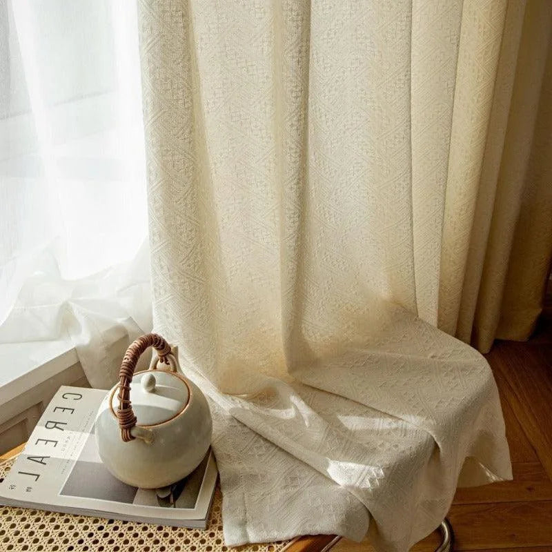 Rideau décoratif beige clair avec motif géométrique, apportant une touche élégante et chaleureuse à la pièce. Idéal pour créer une ambiance lumineuse et raffinée dans le salon. Accessoires de décoration intérieure à proximité, incluant une théière et un magazine.