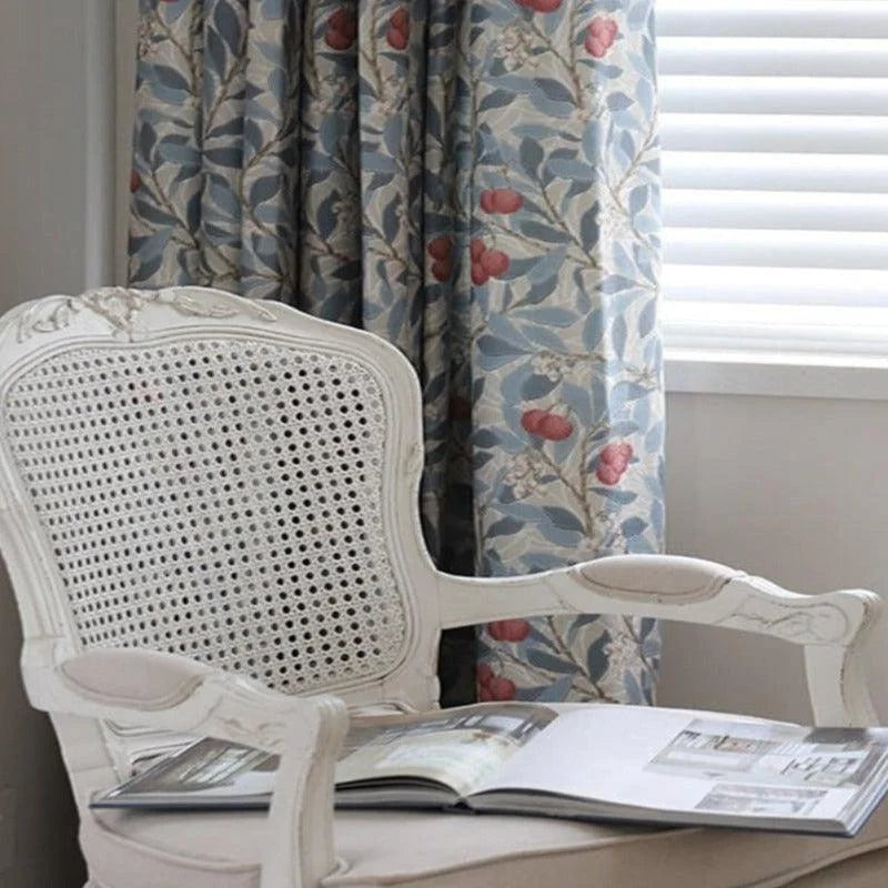 Fauteuil élégant de style vintage avec dossier en cannage, placé dans un intérieur accueillant avec des rideaux fleuris bleus et rouges apportant une touche de charme rustique. Une lumière douce traverse des stores vénitiens, créant une ambiance chaleureuse idéale pour la lecture et la détente.