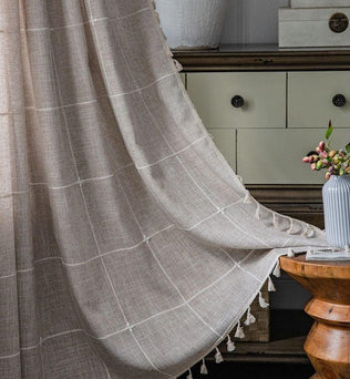 Voilage élégant beige avec motif à carreaux pour une décoration intérieure raffinée. Ce rideau apporte une touche chaleureuse et moderne à votre salon. Il s'intègre parfaitement dans un intérieur chic avec un meuble en bois, mettant en valeur le design d'une table ronde et des accessoires décoratifs comme un vase blanc aux fleurs délicates. Transformez votre espace avec ce rideau au style intemporel, idéal pour filtrer la lumière tout en créant une ambiance conviviale.