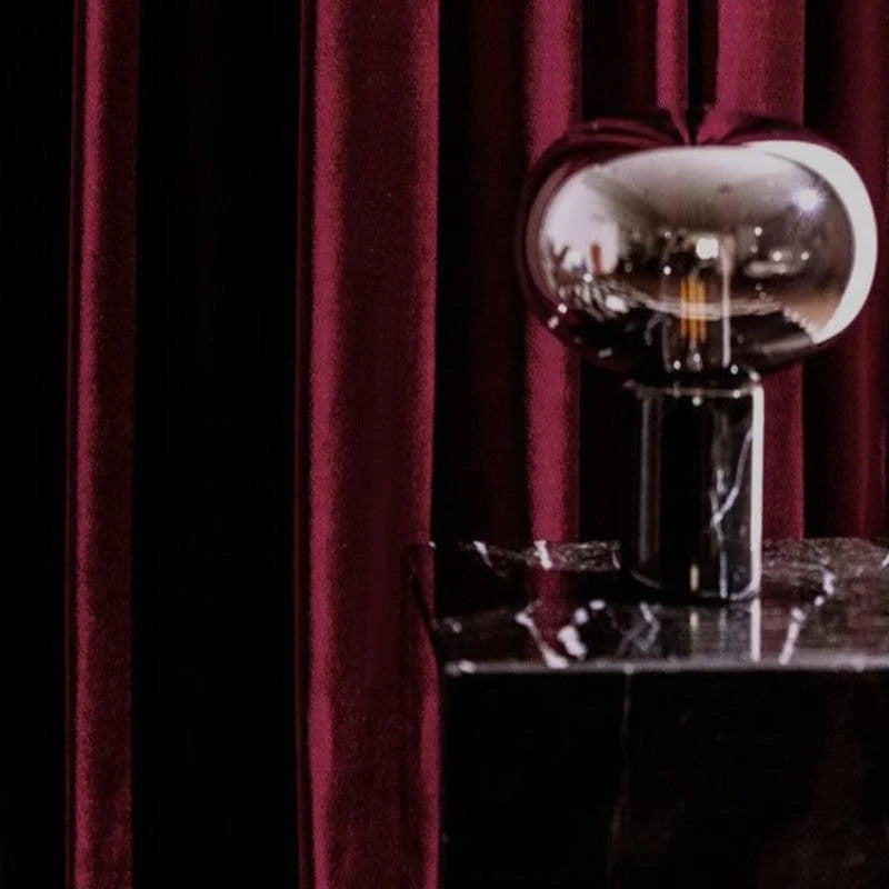 Lampe design moderne posée sur un socle noir élégant, devant des rideaux bordeaux somptueux, créant une ambiance sophistiquée et contemporaine.