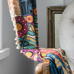 Écharpe multicolore avec motifs abstraits et franges, drapée sur un fauteuil beige dans un intérieur moderne. Accessoire tendance pour rehausser votre style avec des touches vives et artistiques. Parfait pour ajouter une note colorée à votre tenue quotidienne.