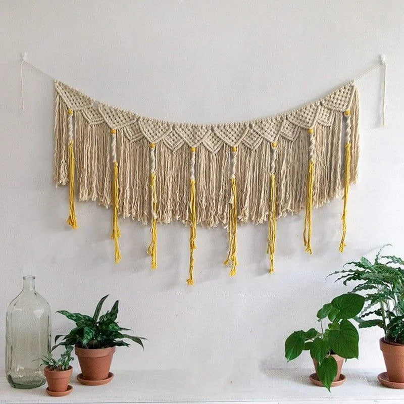 Décoration murale bohème avec franges en macramé suspendue au mur blanc, entourée de plantes vertes en pots. Élément idéal pour un intérieur tendance et chaleureux, ajoutant une touche d'artisanat. Accessoire parfait pour sublimer le salon ou la chambre avec style et élégance.