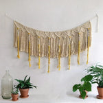 Décoration murale bohème avec franges en macramé suspendue au mur blanc, entourée de plantes vertes en pots. Élément idéal pour un intérieur tendance et chaleureux, ajoutant une touche d'artisanat. Accessoire parfait pour sublimer le salon ou la chambre avec style et élégance.