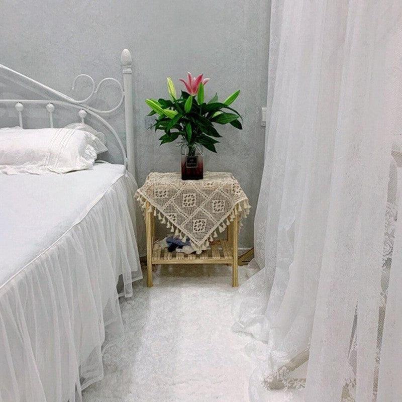 Chambre à coucher élégante avec lit blanc et table d'appoint décorée d'une nappe en crochet. Un vase de fleurs vives ajoute une touche naturelle à l'ambiance apaisante. Rideaux légers encadrent la scène, créant une atmosphère sereine et accueillante. Idéal pour des idées de décoration intérieure.