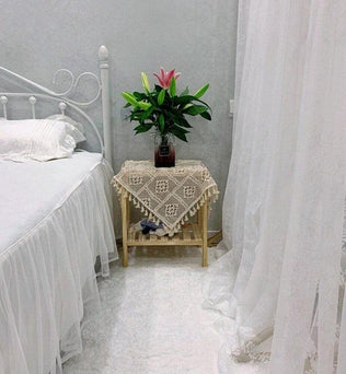 Chambre à coucher élégante avec lit blanc et table d'appoint décorée d'une nappe en crochet. Un vase de fleurs vives ajoute une touche naturelle à l'ambiance apaisante. Rideaux légers encadrent la scène, créant une atmosphère sereine et accueillante. Idéal pour des idées de décoration intérieure.