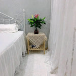 Chambre à coucher élégante avec lit blanc et table d'appoint décorée d'une nappe en crochet. Un vase de fleurs vives ajoute une touche naturelle à l'ambiance apaisante. Rideaux légers encadrent la scène, créant une atmosphère sereine et accueillante. Idéal pour des idées de décoration intérieure.