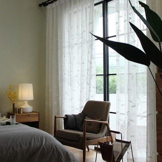 Fauteuil élégant dans une chambre lumineuse avec rideaux translucides, accompagné d'une lampe moderne et d'une plante d'intérieur, créant une ambiance apaisante et contemporaine. Idéal pour un espace de détente.