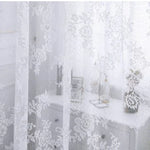 Voilage blanc transparent à motifs floraux habillant une fenêtre devant une coiffeuse, pour une ambiance douce et lumineuse dans la chambre ou le salon.