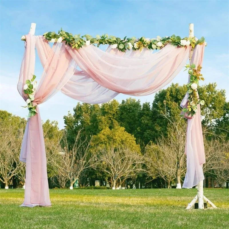 Arche de mariage romantique en plein air avec drapés roses et décorations florales vertes, installée sur une pelouse verte devant des arbres, idéale pour une cérémonie nuptiale élégante et naturelle. Accessoire parfait pour un mariage champêtre ou bohème en extérieur.