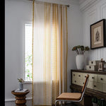 Voilage élégant à motifs géométriques or et blanc, idéal pour une décoration intérieure lumineuse et moderne. Ce rideau apporte une touche sophistiquée à votre salon ou chambre, tout en filtrant délicatement la lumière naturelle. Parfait pour compléter un espace avec des accents boisés et des plantes d'intérieur.