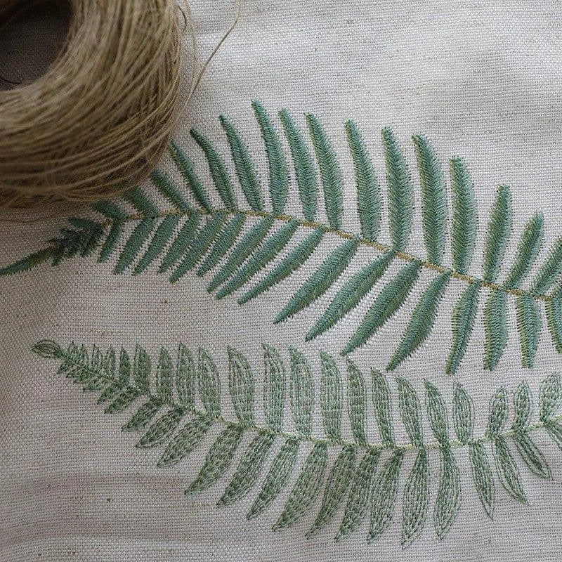 Broderie artisanale avec motif de feuilles de fougère, design élégant et naturel parfait pour décoration intérieure. Image montrant un rouleau de fil à côté de la broderie délicate. Accessoire idéal pour apporter une touche végétale à votre espace.