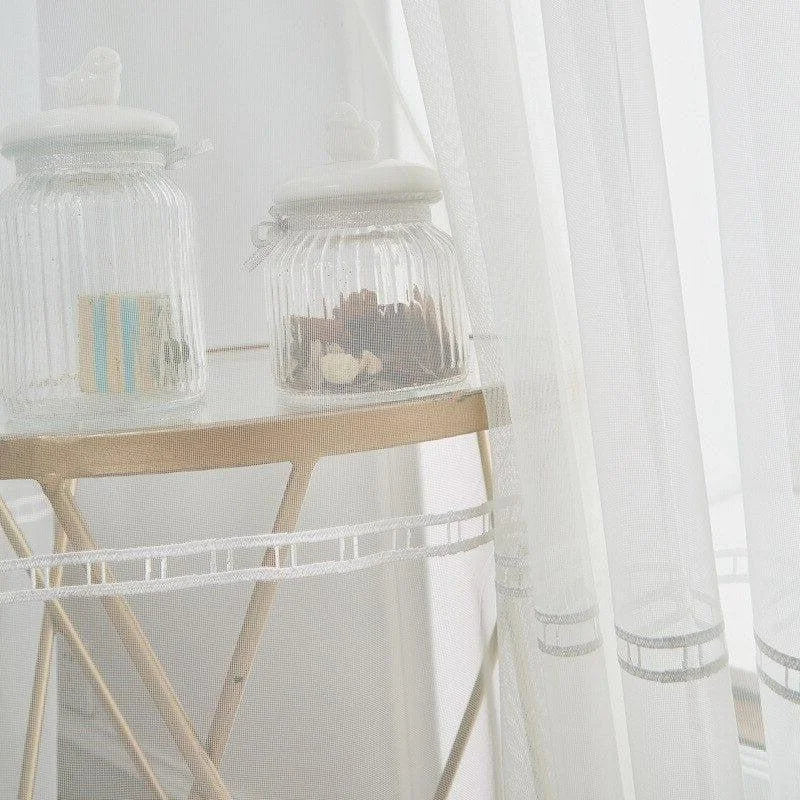 Voilage élégant suspendu à une fenêtre, laissant passer la lumière naturelle dans la pièce avec douceur. Deux bocaux en verre posés sur une table apportent une touche décorative moderne et épurée, idéale pour embellir un intérieur lumineux et raffiné. Atmosphère apaisante et contemporaine.