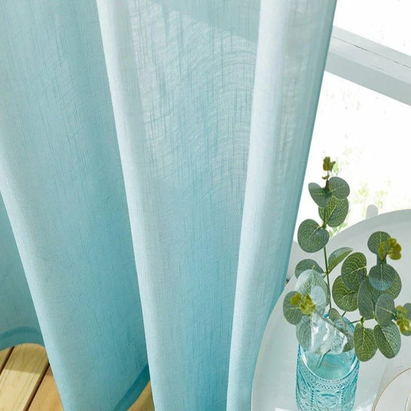 Rideau bleu clair suspendu près d'une fenêtre lumineuse, accompagné d'une plante décorative verte dans un vase sur une table blanche. Idéal pour illuminer les intérieurs et ajouter une touche contemporaine à votre décoration.