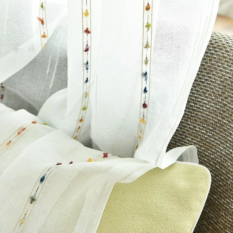 Rideau blanc élégant avec détails brodés colorés suspendu près d'un canapé beige, ajoutant une touche délicate et lumineuse à votre décoration intérieure. Parfait pour créer une ambiance cosy et moderne dans votre salon ou chambre.