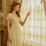 Femme en robe élégante crème touchant un voilage blanc orné de broderies dorées dans un intérieur de style vintage.