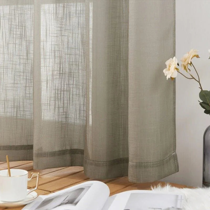 Voilage translucide beige suspendu, offrant une touche élégante et moderne à la décoration intérieure. La lumière douce filtrant à travers le tissu crée une ambiance chaleureuse. Parfait pour le salon ou la chambre, ce rideau s'accompagne d'une tasse de café et d'un livre, suggérant un espace de détente et de lecture. Un vase avec des fleurs fraîches ajoute une note naturelle et apaisante à l'ensemble.