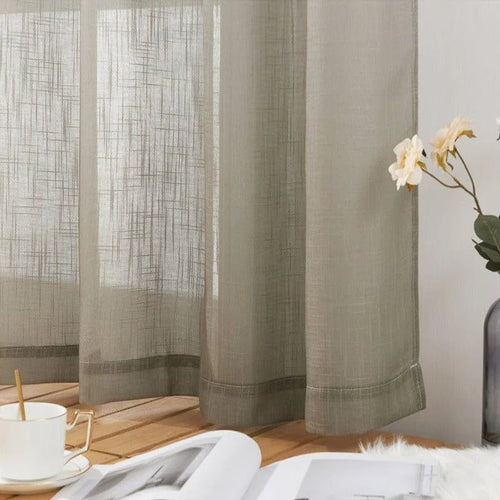 Voilage translucide beige suspendu, offrant une touche élégante et moderne à la décoration intérieure. La lumière douce filtrant à travers le tissu crée une ambiance chaleureuse. Parfait pour le salon ou la chambre, ce rideau s'accompagne d'une tasse de café et d'un livre, suggérant un espace de détente et de lecture. Un vase avec des fleurs fraîches ajoute une note naturelle et apaisante à l'ensemble.