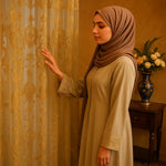Femme arabe en robe beige et hijab marron touchant un voilage doré brodé au style oriental dans une pièce chaleureuse et élégante.