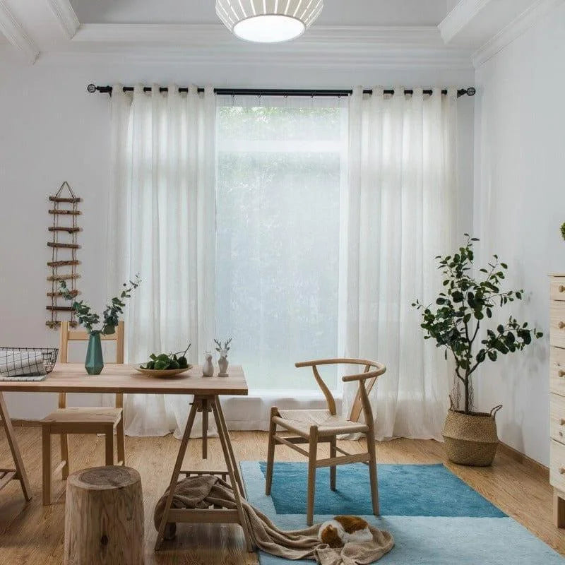 Salle à manger moderne avec une décoration minimaliste, comprenant une table en bois claire entourée de chaises élégantes. Les rideaux légers filtrent la lumière naturelle, créant une atmosphère lumineuse et apaisante. Des plantes d'intérieur ajoutent une touche de verdure, reprenant l'esthétique épurée et naturelle de l'espace. Un tapis bleu apporte une note de couleur, accentuant le style contemporain de la pièce. Idéale pour des moments conviviaux et