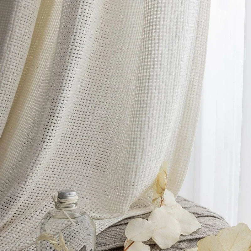 Rideau opaque ivoire avec texture géométrique laissant passer la lumière. Élément de décoration idéal pour embellir un salon ou une chambre à coucher. Flacon en verre et fleurs séchées en arrière-plan pour une ambiance naturelle et apaisante.