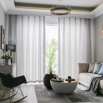 Salon moderne avec rideaux longs et légers laissant passer la lumière naturelle, complété par un canapé confortable et une table basse design, créant une ambiance élégante et accueillante. Décoration raffinée avec un tapis à motifs géométriques et des éléments de décoration contemporains, parfaits pour un intérieur sophistiqué.