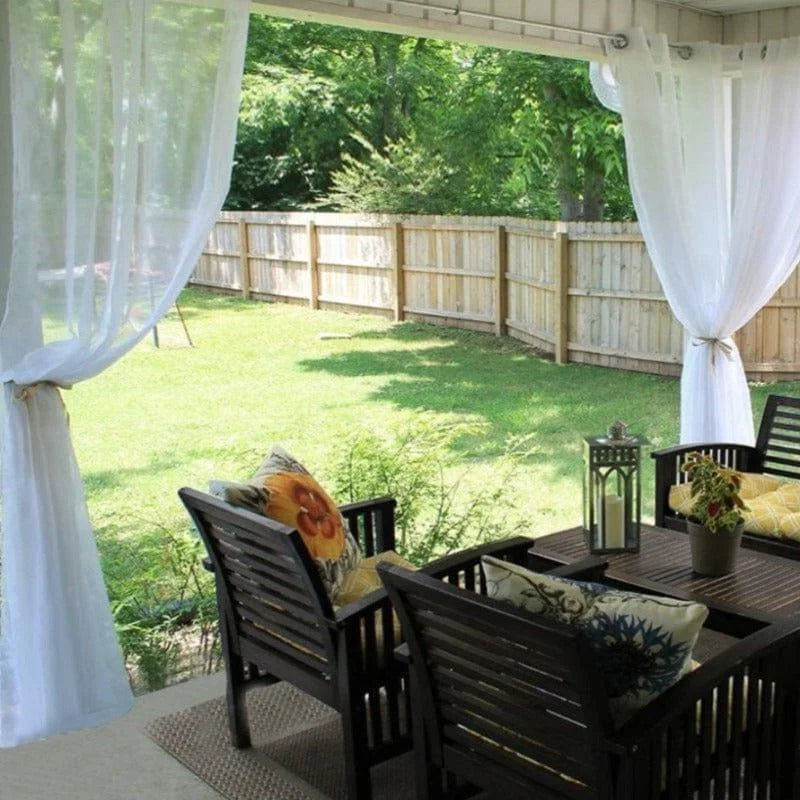 Ensemble de mobilier de jardin comprenant une table et des chaises élégantes disposées sur une terrasse abritée. La vue sur le jardin verdoyant est agrémentée de coussins décoratifs colorés et de lanternes, offrant un espace extérieur confortable et accueillant. Idéal pour des moments de détente ou de convivialité en plein air.
