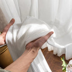 Main tenant un rideau blanc avec motif subtil, soulignant sa légèreté et sa qualité supérieure, parfait pour apporter une touche élégante et aérée à votre intérieur. Luminosité amplifiée grâce à sa texture soignée. Idéal pour salon ou chambre, offrant une ambiance chaleureuse et raffinée.