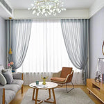 Salon moderne avec décoration élégante, comprenant un canapé confortable, un fauteuil design et une table basse en bois. Grands rideaux bleus encadrant une fenêtre lumineuse, créant une ambiance chaleureuse et accueillante. Lustre suspendu au plafond apportant une touche contemporaine à l'ensemble. Meuble de rangement en bois clair avec téléviseur moderne, idéal pour un espace de vie harmonieux et chic.