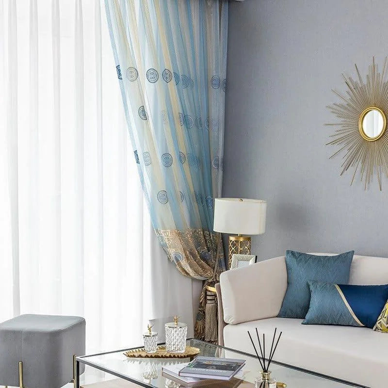 Salon moderne et élégant avec des rideaux bleus décoratifs, un canapé blanc agrémenté de coussins bleus, une table basse en verre ornée d'accessoires sophistiqués, et une lampe de table raffinée, créant une atmosphère chic et apaisante. Décoration murale en forme de soleil doré apportant une touche de style unique.